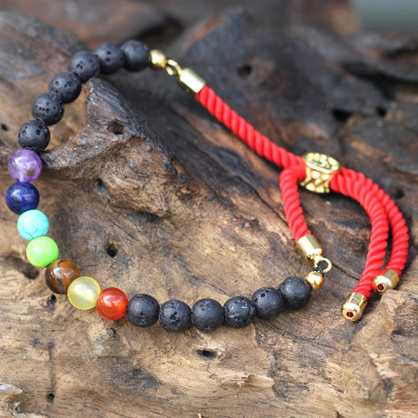 A 18K gold plated red string bracelet with lava stone chakra beads on a wooden surface.