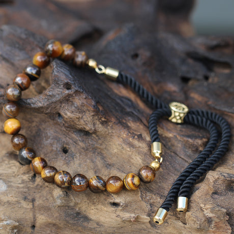 A gold-plated bracelet with black string detailing and Tiger's Eye beads, displayed on a wooden surface.