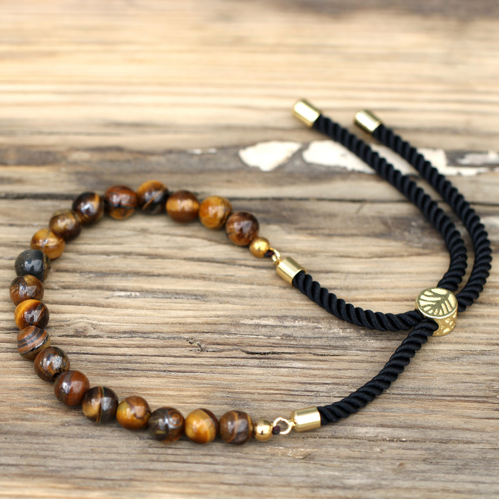 A gold-plated bracelet with black string detailing and Tiger's Eye beads, displayed on a wooden surface.