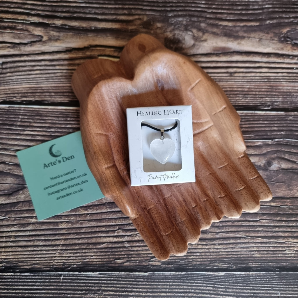 A white crystal heart on a black cord necklace inside a white display box, displayed on a pair of wooden hands