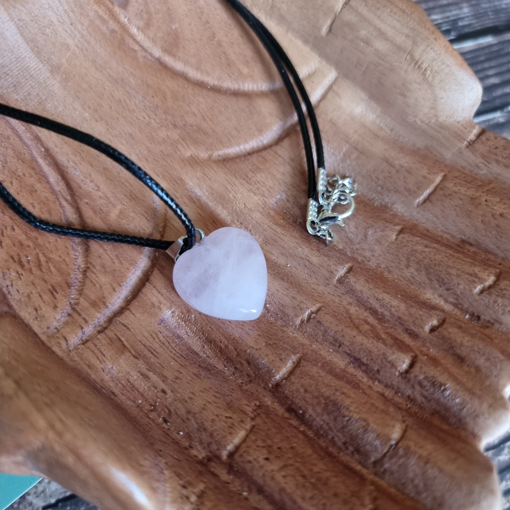 A pink crystal heart on a black cord necklace displayed on a pair of wooden hands
