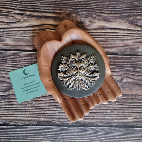 A bronze-effect green man incense holder made of resin, displayed on a pair of wooden hands on a wooden surface next to a green Arte's Den business card. 