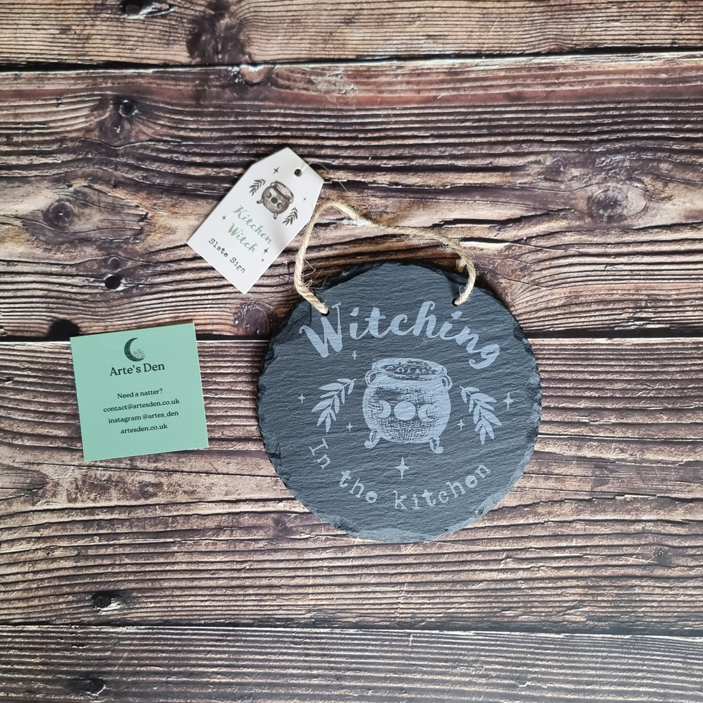 A circular slate sign with the words 'Witching in the Kitchen' on a wooden surface