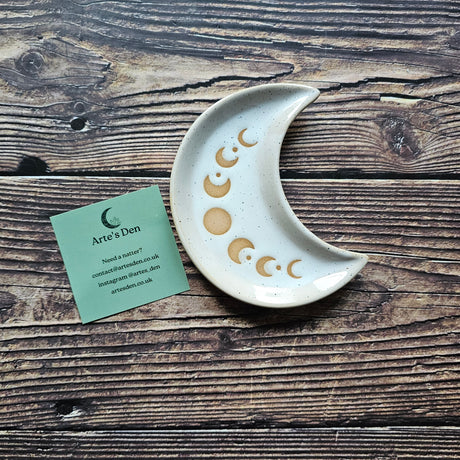 A white, speckled crescent moon trinket dish with golden moon phase detailing on a wooden surface. 