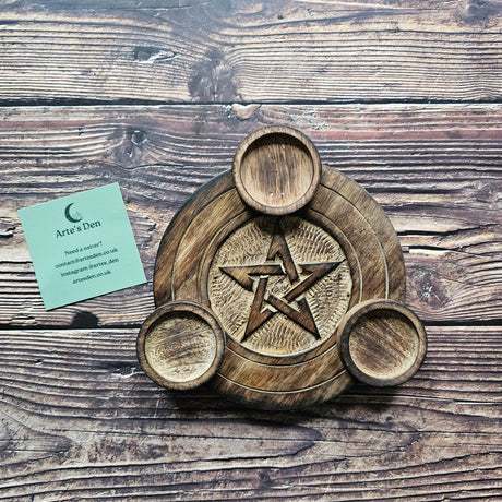 A round wooden tea light holder with an engraved pentagram design with space for three tea lights. Sits against a wooden background with a green Arte's Den business card