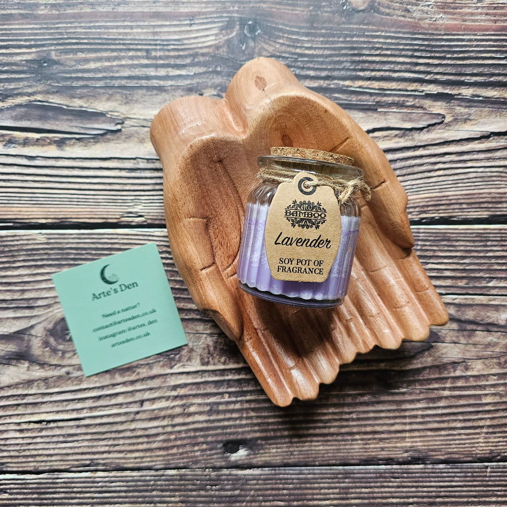 A purple soy candle inside a glass jar with a cork lid, adorned with a brown label that reads 'Lavender SOY POT OF FRAGRANCE'. Laid on top of a pair of wooden hands. 