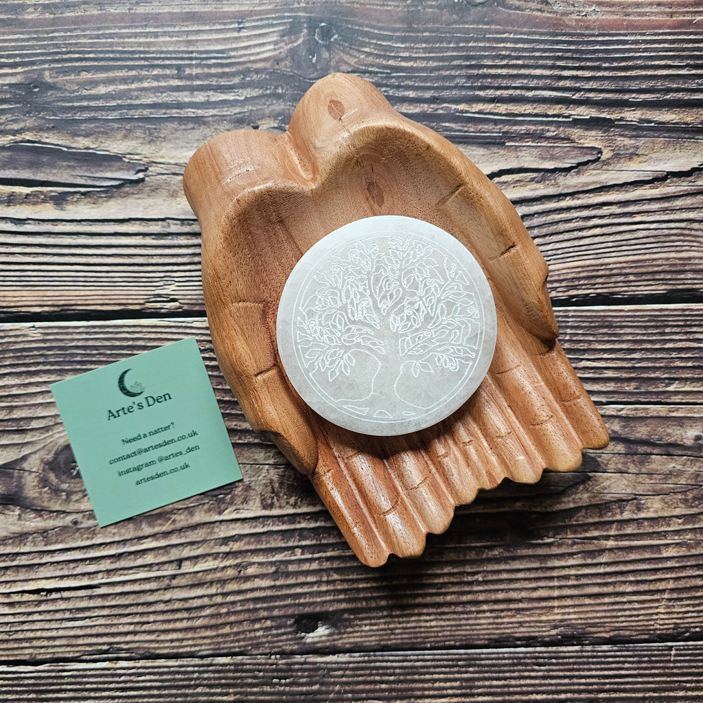 A round selenite crystal charging plate with an engraved Tree of Life design, placed on a wooden surface.