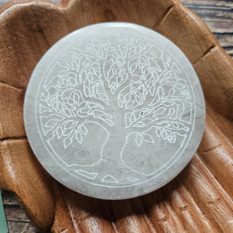 A round selenite crystal charging plate with an engraved Tree of Life design, placed on a wooden surface.