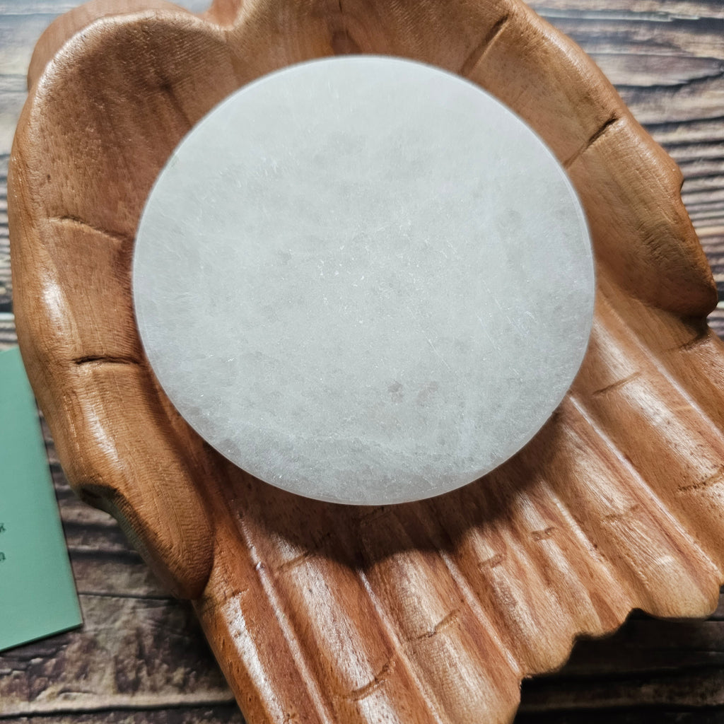 A round selenite crystal charging plate, placed on a wooden surface.
