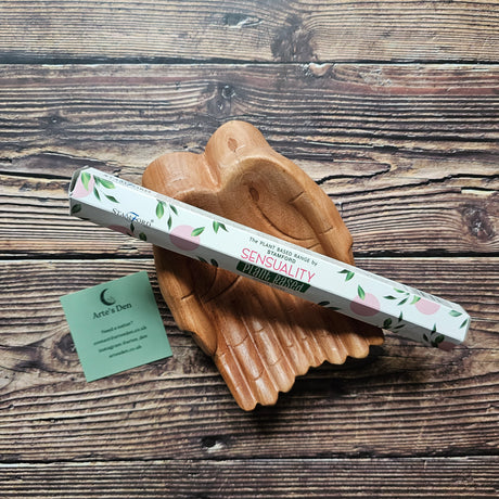 "Sensuality" incense sticks in white packaging with green leaves, placed on a pair of wooden hands on a wooden surface.