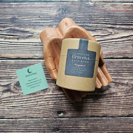 A brown package with black label stating "gemstone bracelet bath bomb" which is laid on a pair of wooden hands on a wooden surface. 
