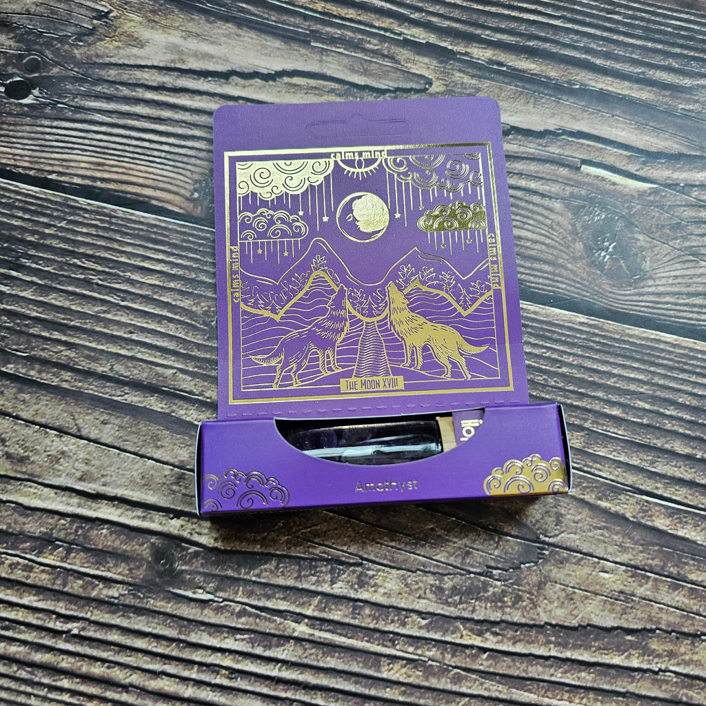 A purple roll-on essential oil bottle with a gold-printed tarot card, set against a packaging with a wolf illustration, laid on a wooden surface. 