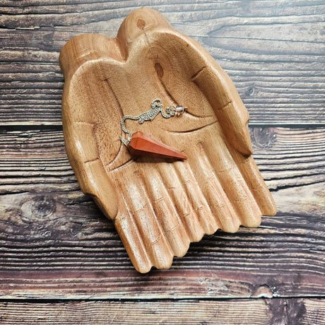 A red jasper pendulum with a silver chain, placed in a wooden hand-shaped dish, on a wooden background.