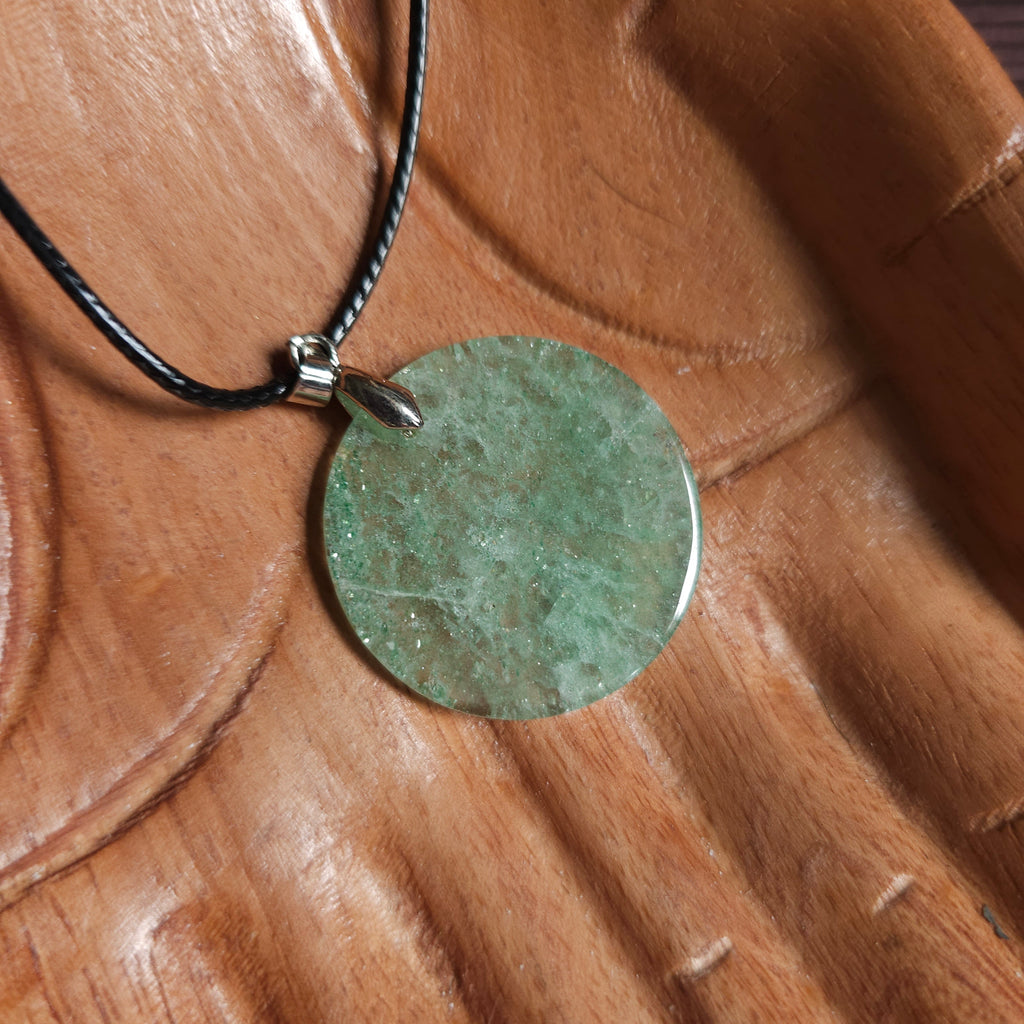 A coin-shaped crystal green aventurine pendant, attached to a black cord, displayed on a wooden pair of hands