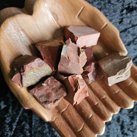 A collection of rough, opaque, deep Red Jasper stones with black and white veins, placed in a wooden hand bowl on a dark background.