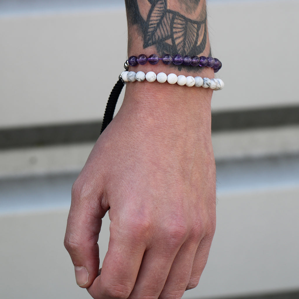 A silver-plated bracelet with navy string detailing and white howlite beads, worn on a tattooed wrist next to an Amethyst beaded bracelet. 