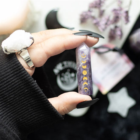 A hand holding a Amethyst crystal point with gold-tone moon phase engraving, accompanied by a matching drawstring bag.