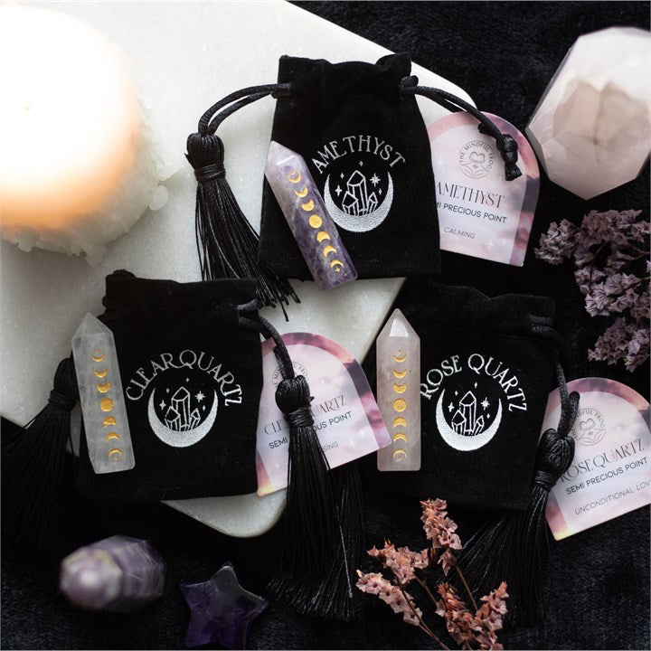 Three crystal points; one Amethyst, one Rose Quartz and one Clear Quartz, all with gold-tone moon phase engraving sit on top of a black velvet bag which states the name of each crystal.
