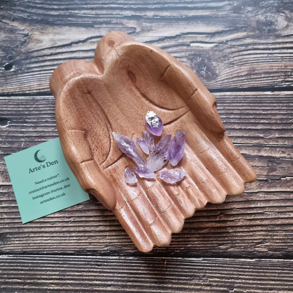 A collection of small pointed Amethyst crystals displayed in a pair of wooden hands on a wooden surface next to a green Arte's Den business card