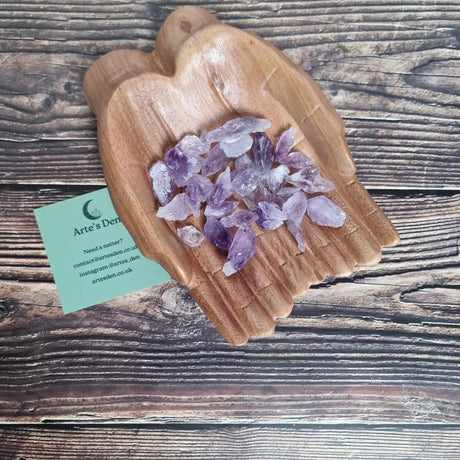 A collection of small pointed Amethyst crystals displayed in a pair of wooden hands on a wooden surface next to a green Arte's Den business card