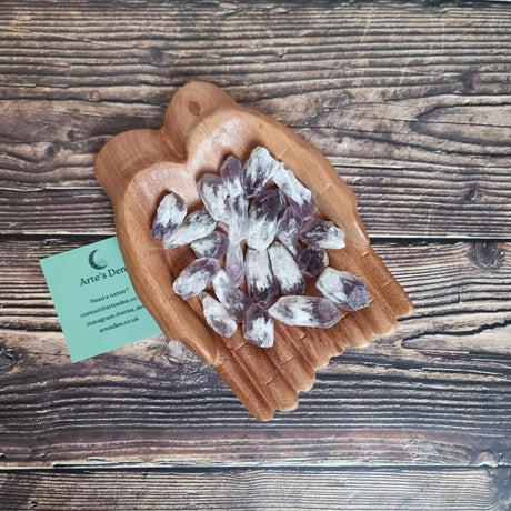 A collection of rough pointed Amethyst crystals displayed in a pair of wooden hands, on a wooden surface with a business card labelled 'Arte's Den' on the side.