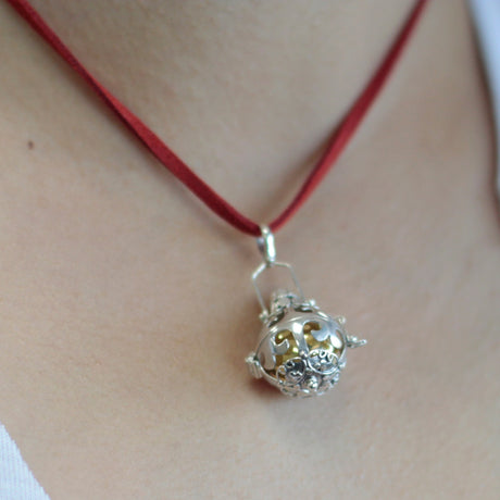 A silver angel bell necklace with a red cord and silver detailing on the necklace and bell.