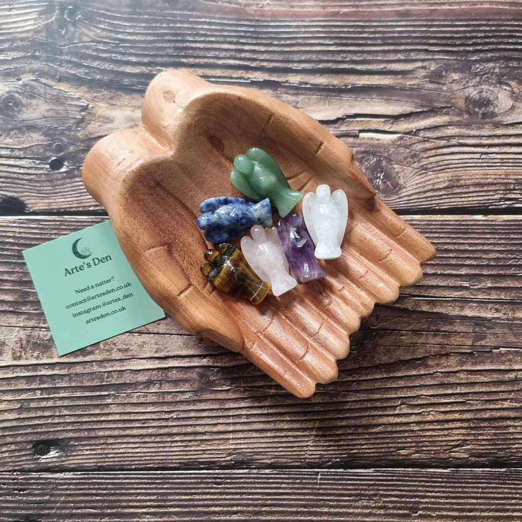 A collection of crystal healing angels in various colours including purple, clear, green, and brown, displayed on a pair of wooden hands on a wooden surface next to a green Arte's Den business card 