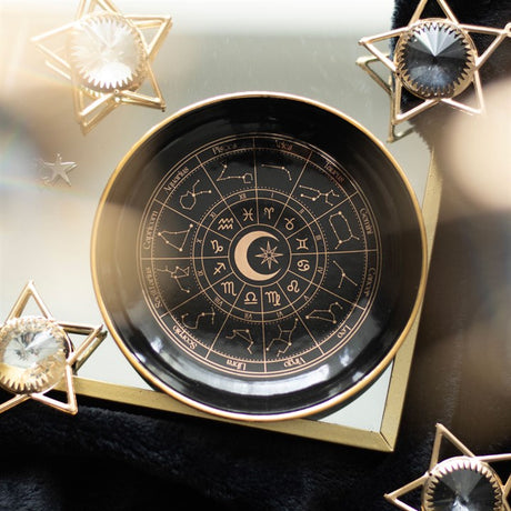 A black trinket dish with a gold effect astrology wheel design, displayed on a surface with star-shaped embellishments.