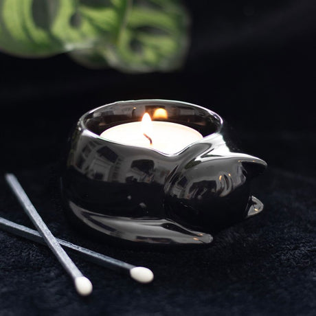 A black cat-shaped tea light candle holder with a lit candle inside, placed on a dark surface with some decorative greenery in the background.