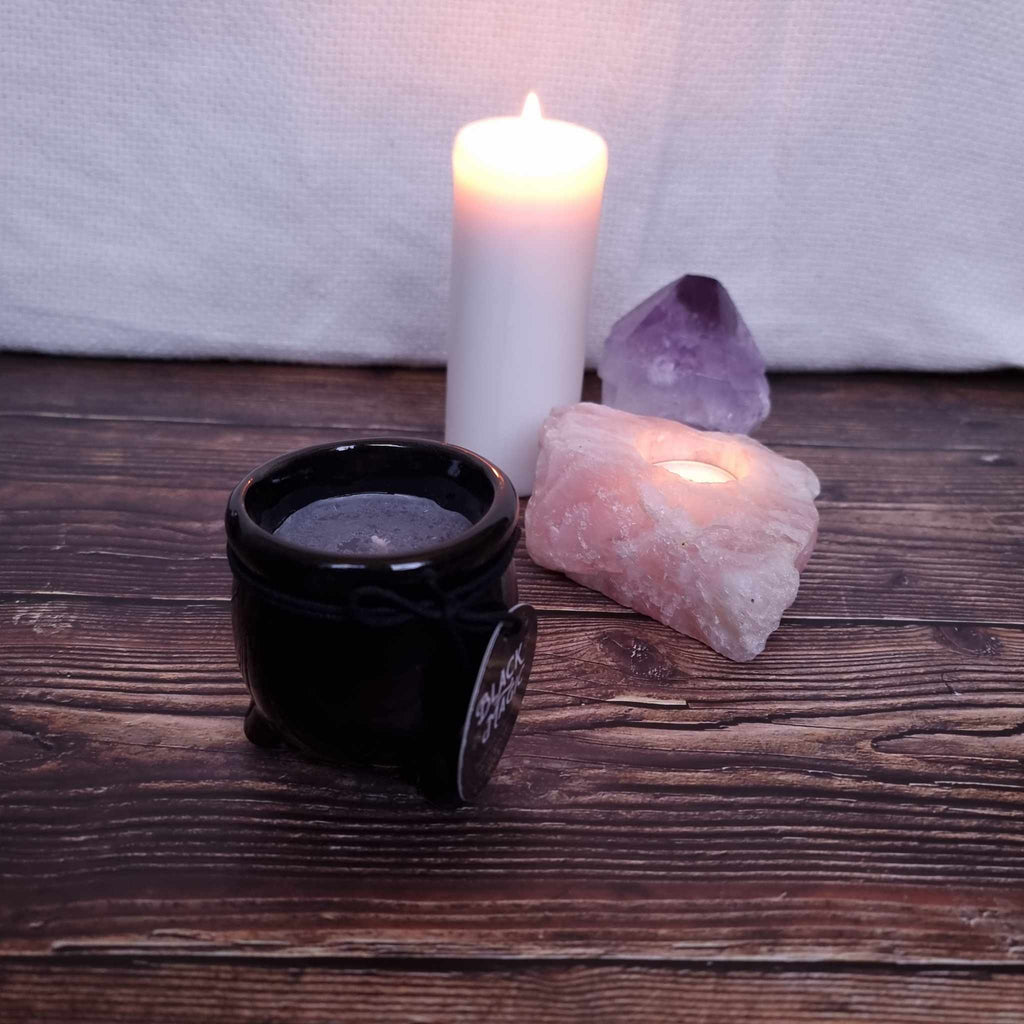 A black cauldron-shaped candle with dark wax and with a label indicating the scent and 'Black Magic' branding. Sits in front of a lit pillar candle and two crystals on a wooden surface. 
