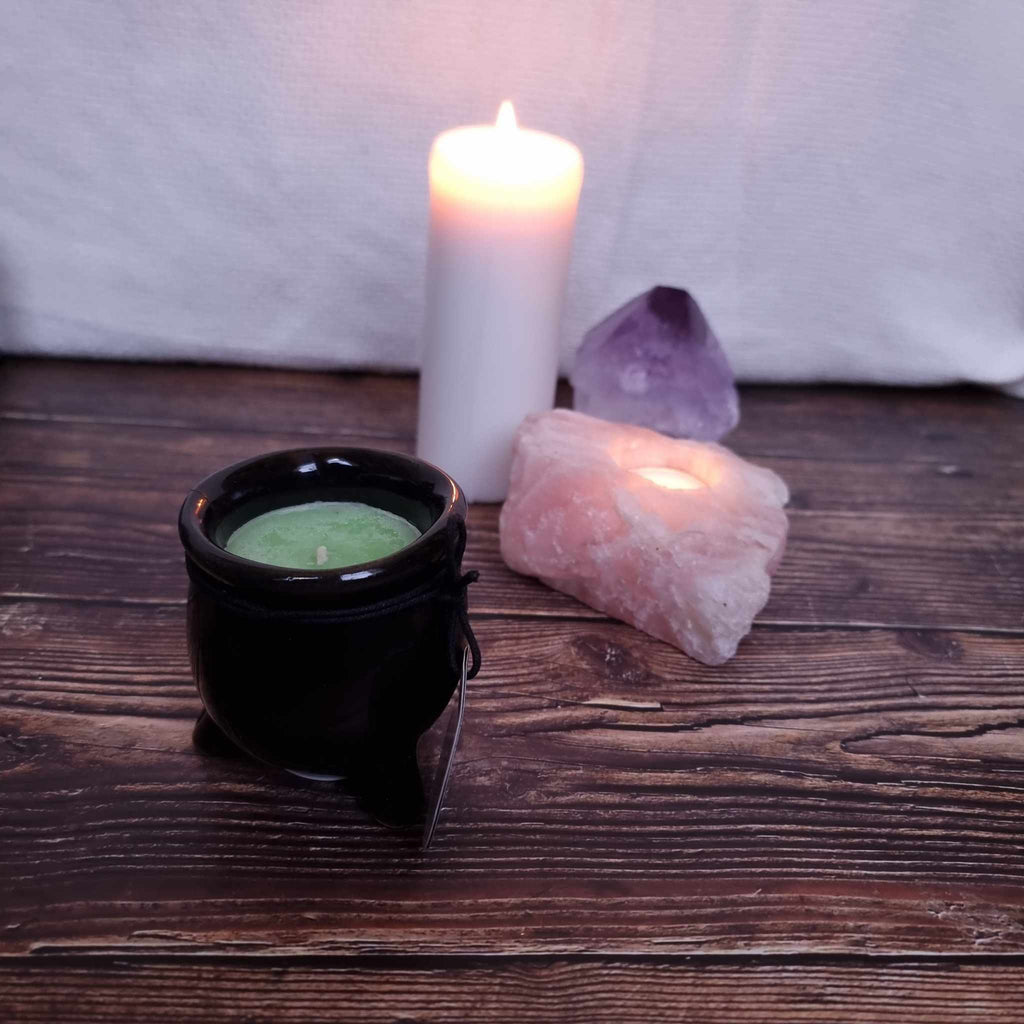 A black cauldron-shaped candle with green wax and with a label indicating the scent and 'Black Magic' branding. Sits in front of a lit pillar candle and two crystals on a wooden surface. 