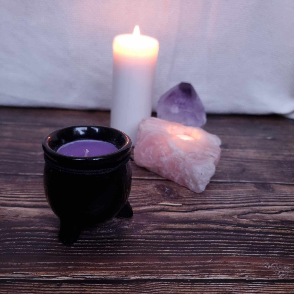 A black cauldron-shaped candle with purple wax and with a label indicating the scent and 'Black Magic' branding. Sits in front of a lit pillar candle and two crystals on a wooden surface. 