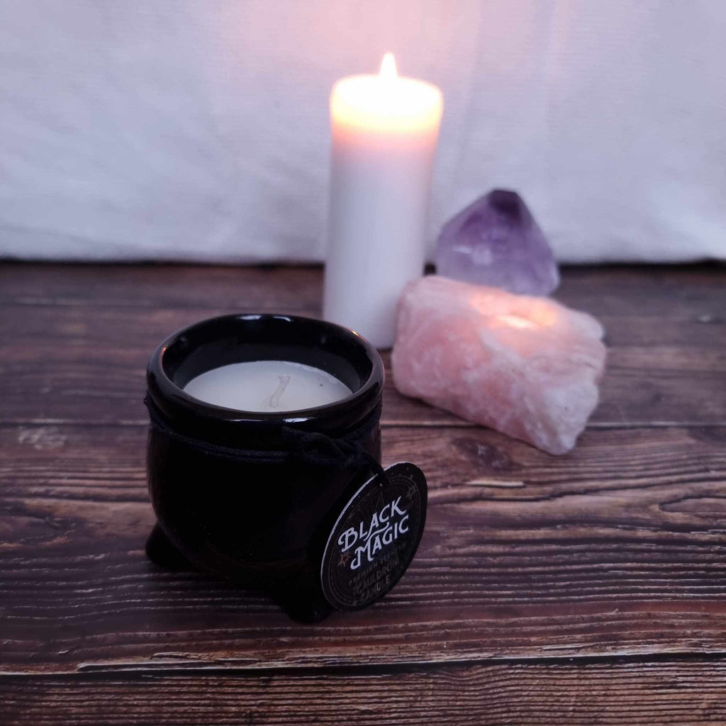 A black cauldron-shaped candle with white wax and with a label indicating the scent and 'Black Magic' branding. Sits in front of a lit pillar candle and two crystals on a wooden surface. 