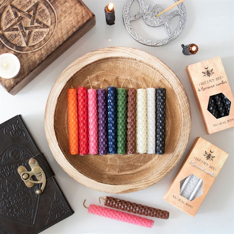 A selection of beeswax candles in various colours, displayed in a round wooden bowl, with a book and other magical items in the background.