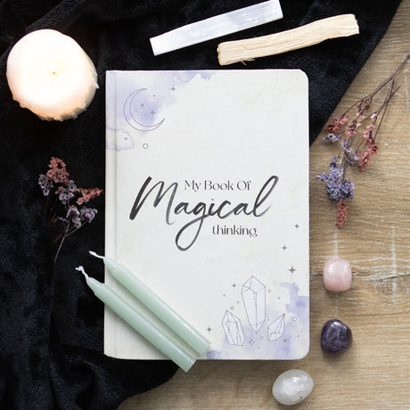 An A5 "My Book of Magical Thinking" notebook with crystal and moon design on a wooden and black fabric background with crystals, candles and dried flowers 