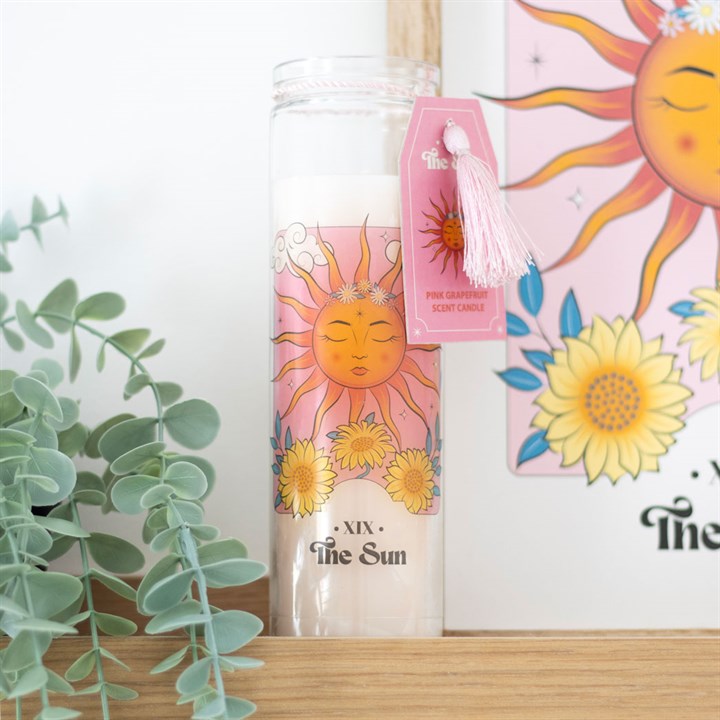 A tube candle with a sun design, featuring an orange and pink colour scheme and a decorative tassel, placed on a shelf with a plant and a framed artwork in the background.