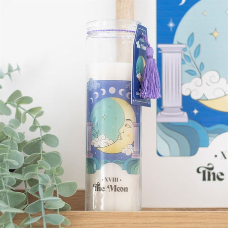 A tube candle with a moon design, featuring a blue colour scheme and a decorative tassel, placed on a shelf with a plant and a framed artwork in the background.