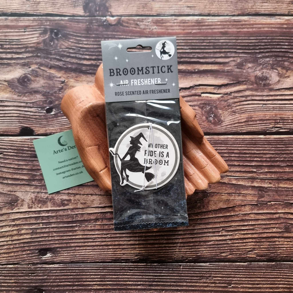 A pair of wooden hands holding a packaged air freshener with a moon and witch design and rose scented description. It is laid on a wooden background next to a green Arte's Den business card.