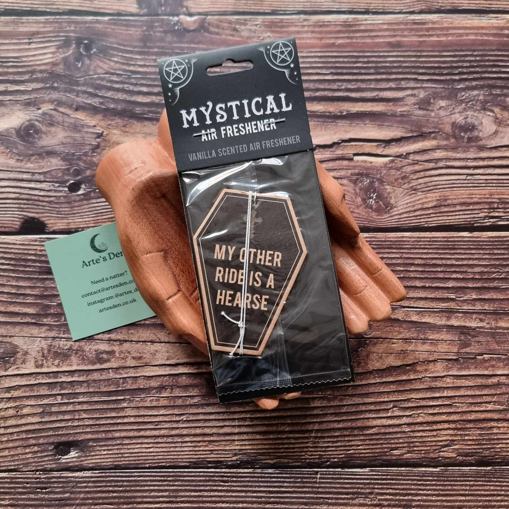 A pair of wooden hands holding a packaged air freshener with a coffin design and vanilla scented description. It is laid on a wooden background next to a green Arte's Den business card.