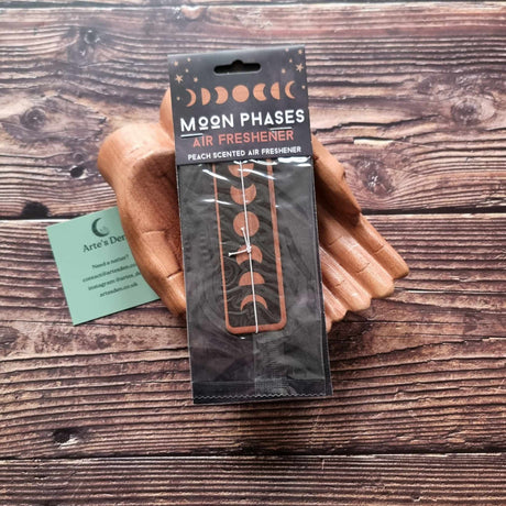 A pair of wooden hands holding a packaged air freshener with a moon phases design and peach scented description. It is laid on a wooden background next to a green Arte's Den business card.