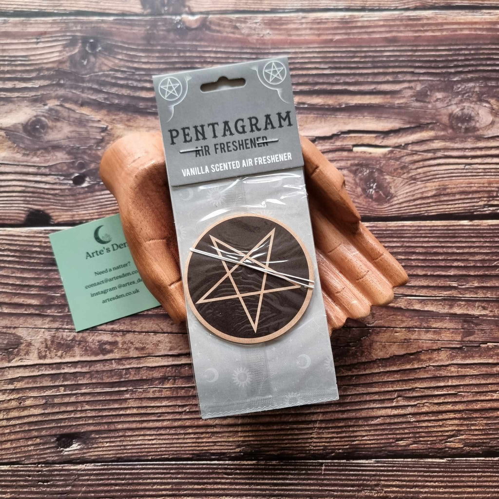 A pair of wooden hands holding a packaged air freshener with a pentagram design and vanilla scented description. It is laid on a wooden background next to a green Arte's Den business card.