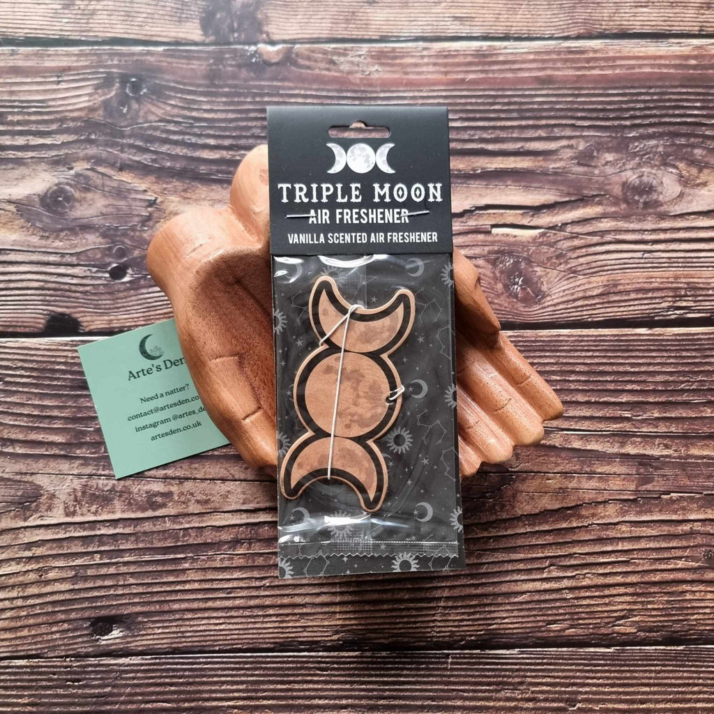 A pair of wooden hands holding a packaged air freshener with a triple moon design and vanilla scented description. It is laid on a wooden background next to a green Arte's Den business card.