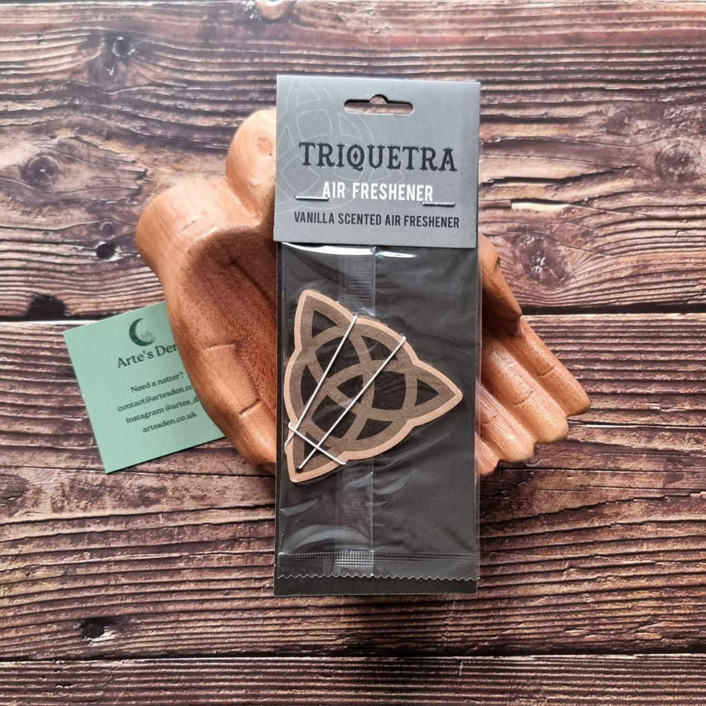 A pair of wooden hands holding a packaged air freshener with a triquetra design and vanilla scented description. It is laid on a wooden background next to a green Arte's Den business card.