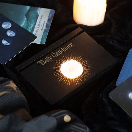 A black tarot and oracle card stand with "Daily Guidance" and a lit tea light in the centre, displayed on a dark background.