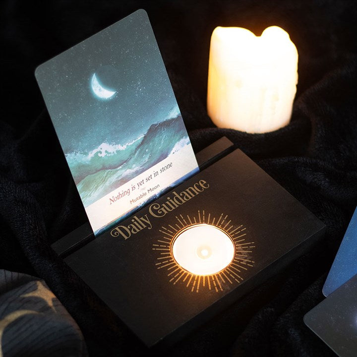A black tarot and oracle card stand with "Daily Guidance" and a lit tea light in the centre and a card, displayed on a dark background.