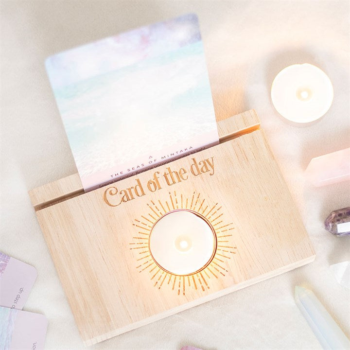 A wooden tarot and oracle card stand with "Card of the day" and a tea light in the centre and a card, displayed on a light background.