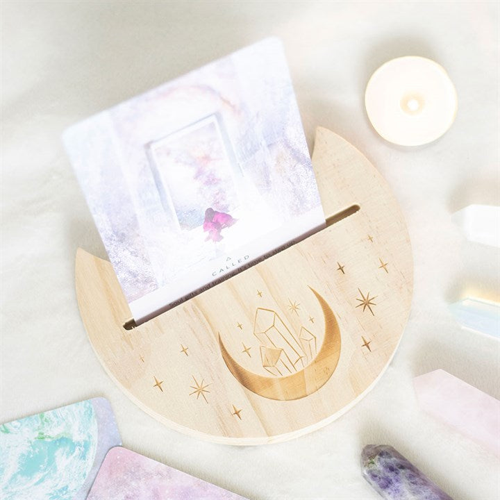 A wooden crescent moon shaped tarot and oracle card stand with a moon and crystal design and a card, displayed on a light background.