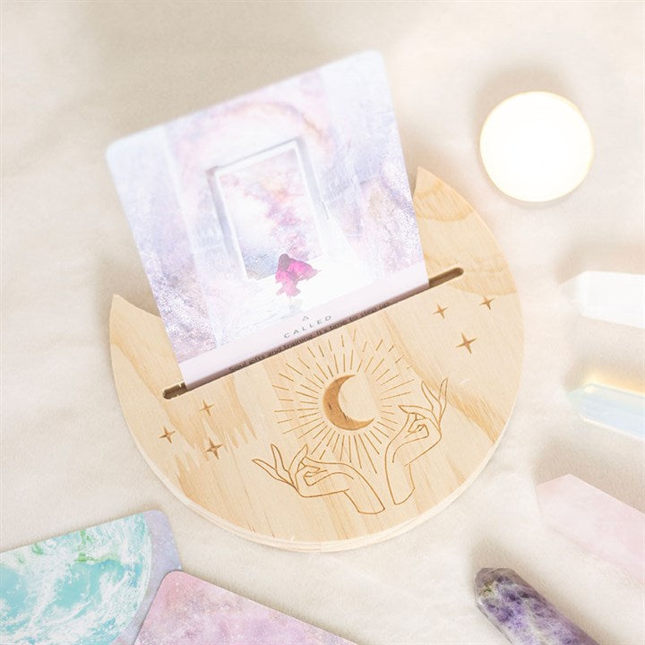A wooden crescent moon shaped tarot and oracle card stand with a moon and hand design and a card, displayed on a light background.