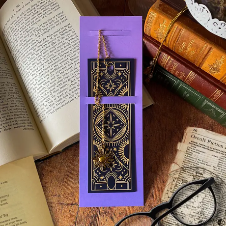 Purple packaging with a golden designed bookmark on a wooden surface with books and glasses.