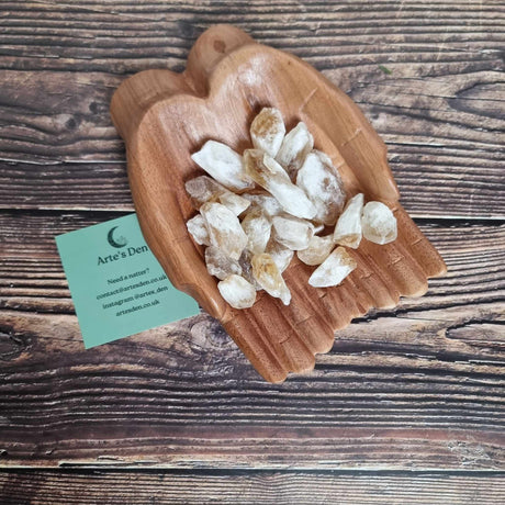 A collection of pointed citrine topaz crystals displayed in a pair of wooden hands on a wooden surface next to a green Arte's Den business card